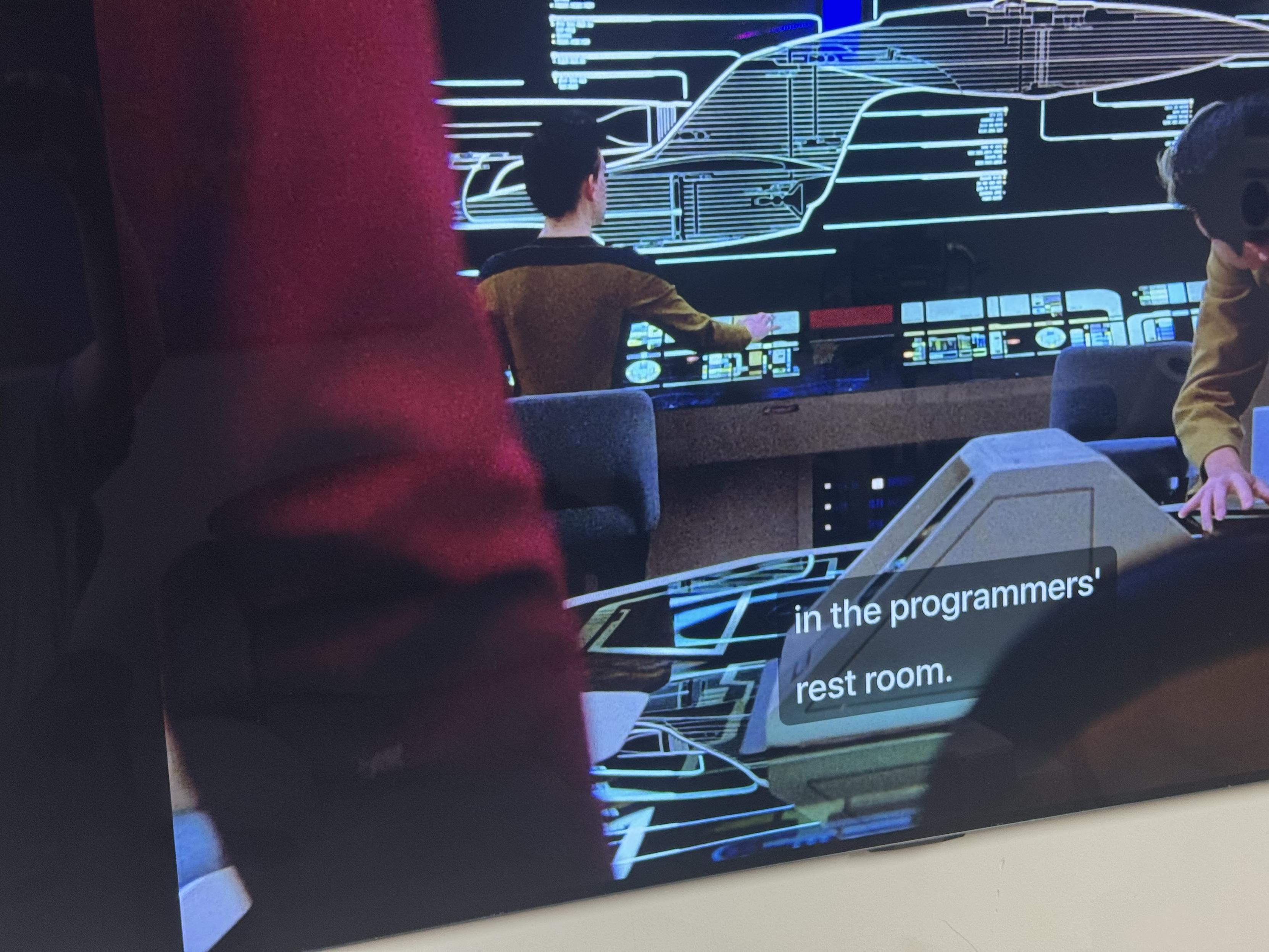 An interior scene from a sci-fi setting, featuring two individuals interacting with a futuristic control panel and a digital display of a starship design. The environment appears to resemble a command room or programmers' workspace. An interior scene from a sci-fi setting, featuring two individuals interacting with a futuristic control panel and a digital display of a starship design. The environment appears to resemble a command room or programmers' workspace.