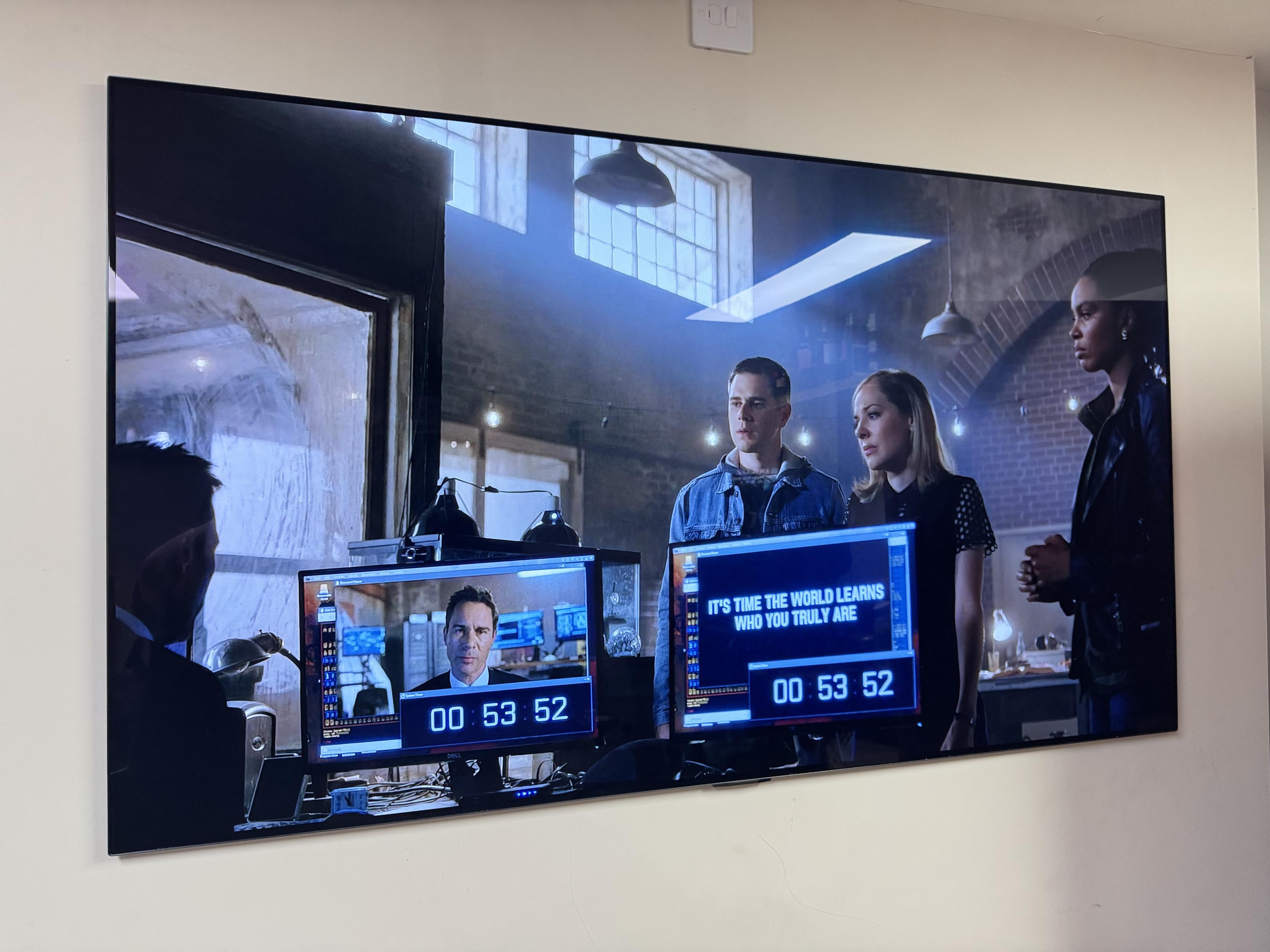 The image shows a scene from a film or TV show featuring four characters in a modern office environment. Two monitors display a countdown timer and a message, "IT'S TIME THE WORLD LEARNS WHO YOU TRULY ARE”.
